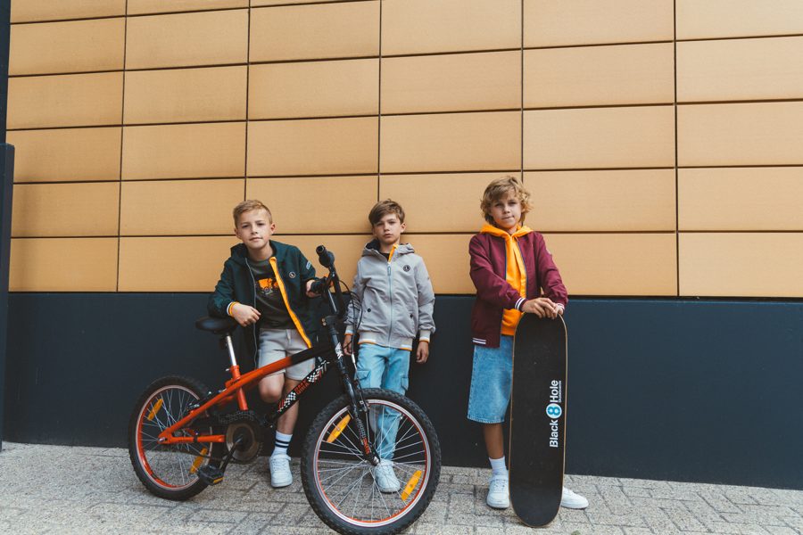Drie kinderen met fiets en skateboard voor een gevel, in kleding van Tygo & Vito